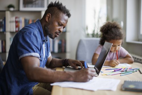 Father working on laptop while his daughter drawing beside him at home